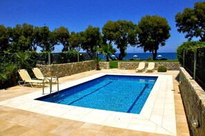 A rectangular swimming pool with a stone wall and tiled deck, surrounded by lounge chairs and a view of the sea.