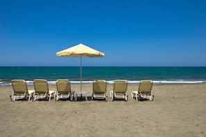 Une rangée de chaises de plage et un seul parasol sur une plage de sable, avec un ciel bleu dégagé et l’océan en arrière-plan.