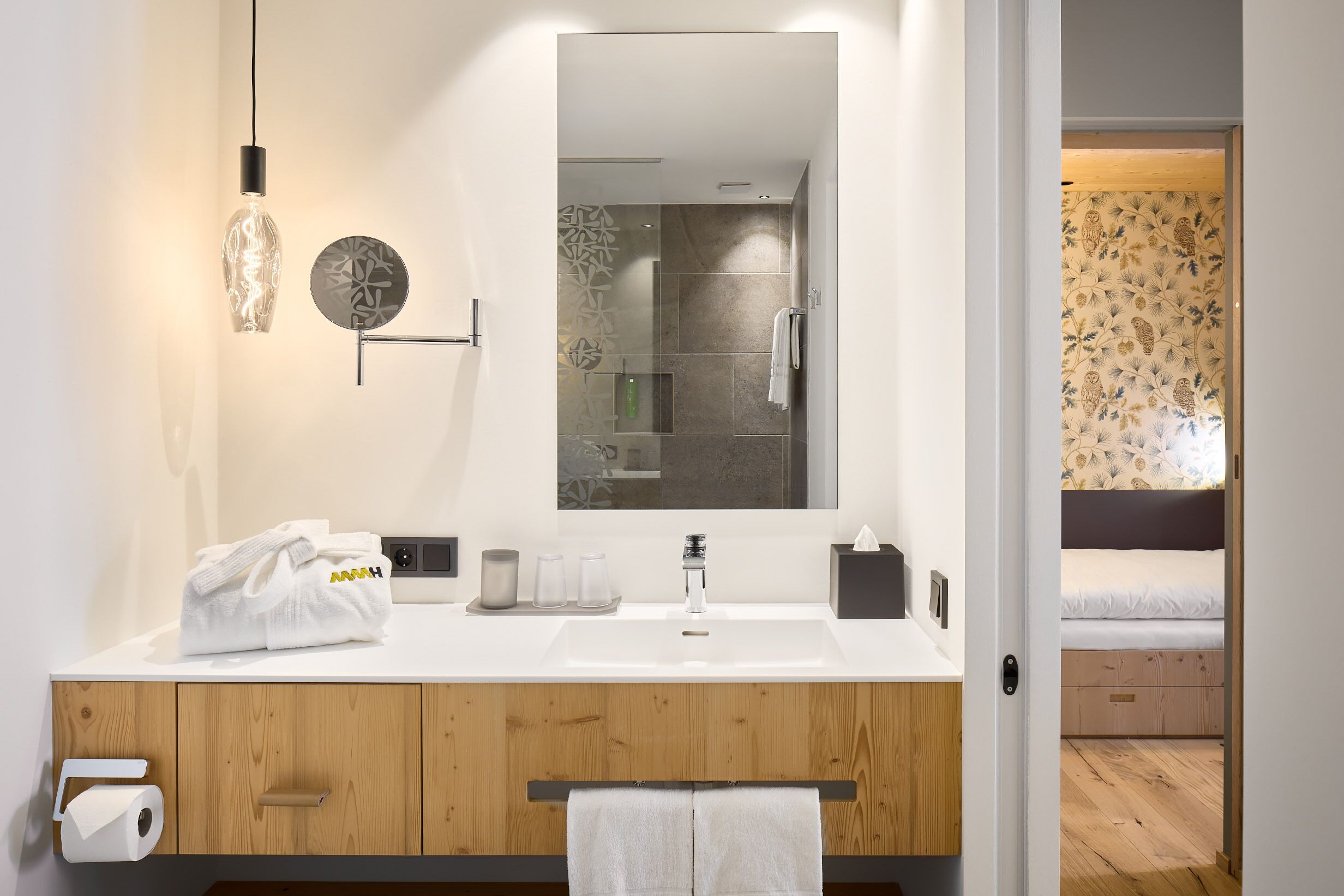A modern bathroom with a white sink, wooden vanity, and a large mirror. There is a towel rack with a white towel, a glass bottle, and a decorative plate on the vanity.
