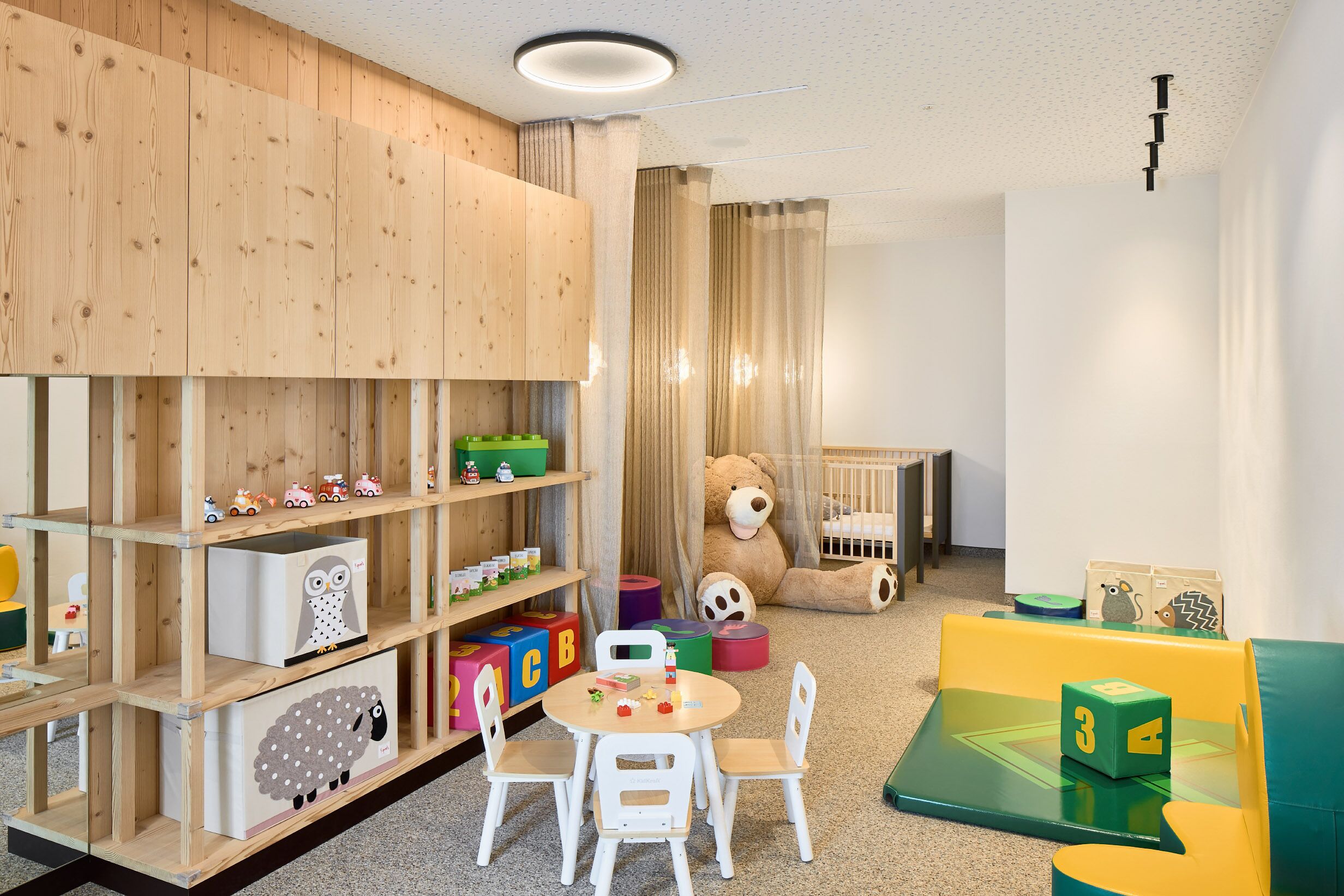 Une salle de jeux pour enfants avec des étagères en bois, un grand ours en peluche et des jouets colorés.