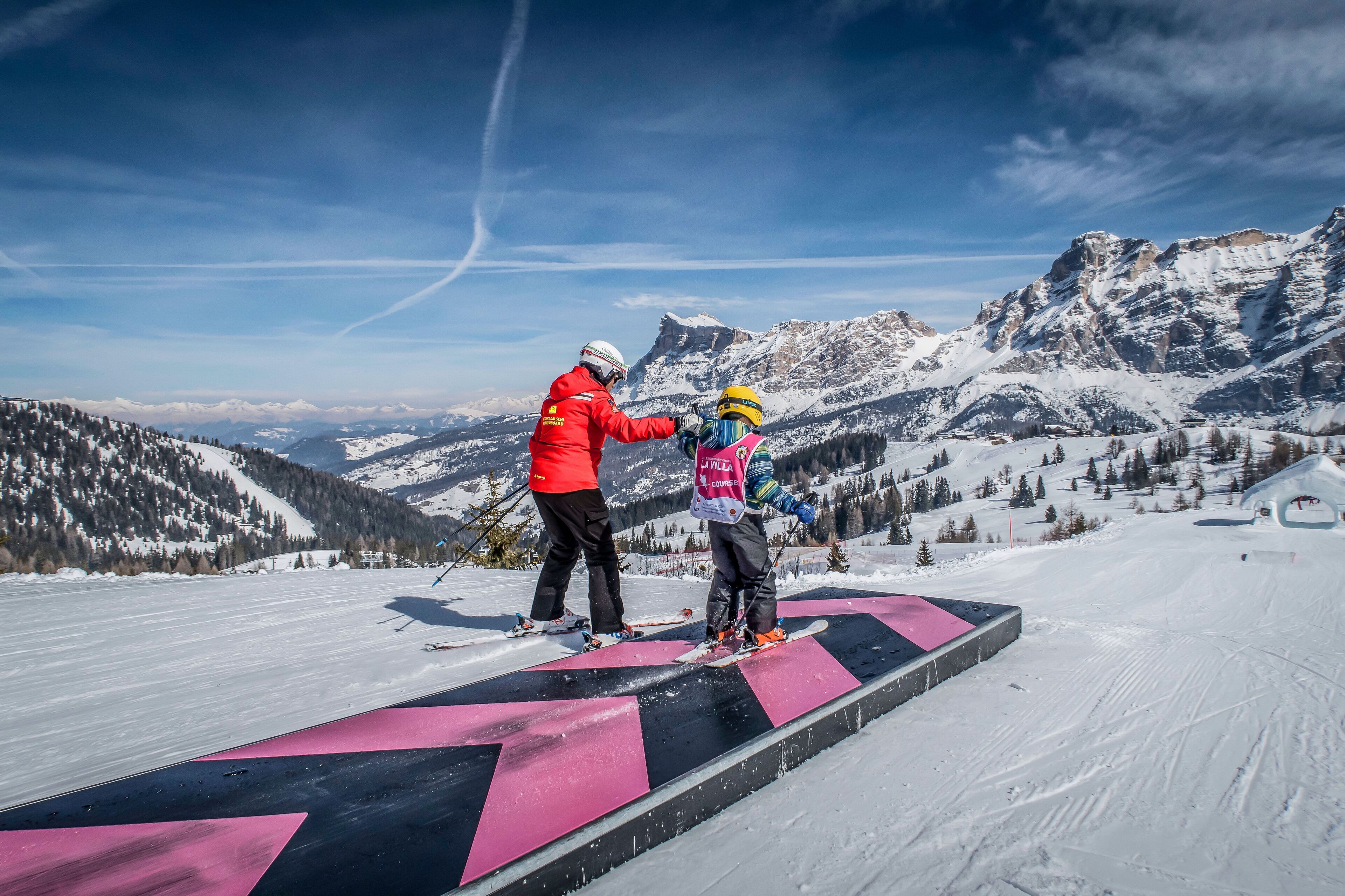 Een skipiste met een persoon in een rode jas en een ander in een blauwe jas, staand op een platform met een roze en zwart ontwerp.