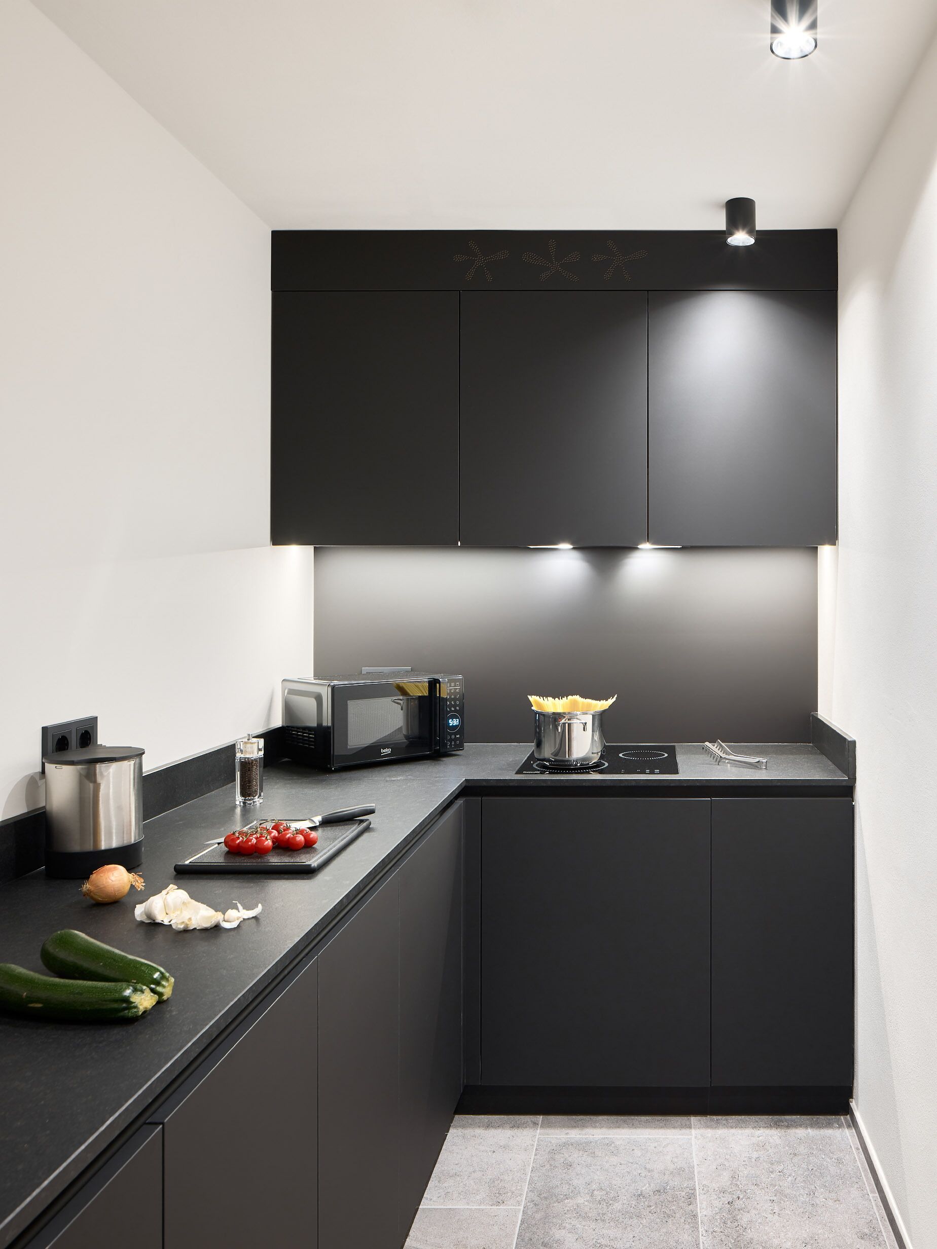A modern kitchen with dark cabinetry, a microwave, a kettle, and fresh vegetables on the counter.
