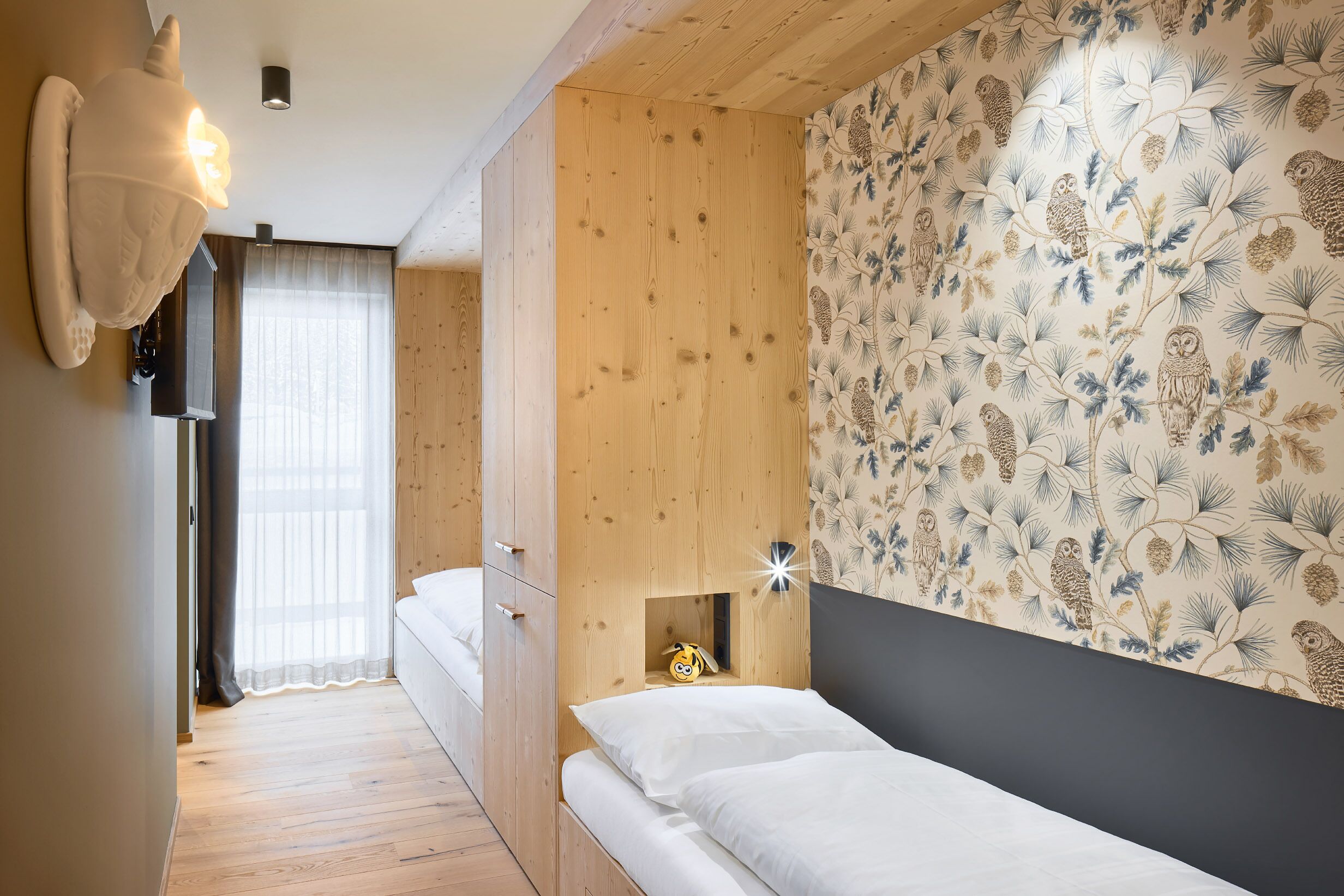 A bedroom with a wooden wardrobe, a bed with white bedding, a wall-mounted light fixture, and a floral wallpaper.