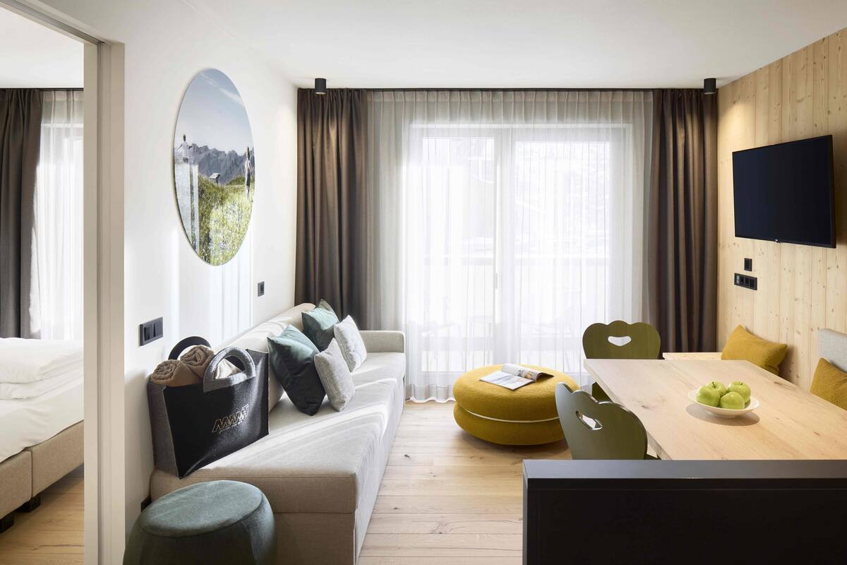 A modern hotel room with a sofa, ottoman, dining table, and a flat-screen TV.