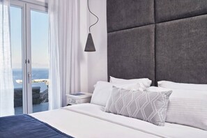 Ein modernes Schlafzimmer mit einem großen Bett, einem Nachttisch, einer Nachttischlampe und Blick auf das Meer durch eine Glastür.
