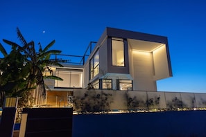 A modern two-story house with large windows and a balcony, surrounded by a well-lit garden.