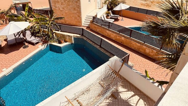 Zona de piscina amb vista despejada al mar, un edifici de façana de pedra i una platja sorrenca al fons.