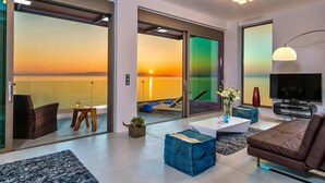 A modern living room with a large sliding glass door leading to a balcony with a sunset view. - Villa Blue Lagoon -Unifying your luxury senses -SEAFRONT (Chania Akrotiri)