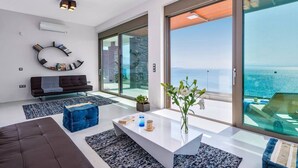 A modern living room with a large glass balcony overlooking the sea, a white coffee table, and a black sofa. - Villa Blue Lagoon -Unifying your luxury senses -SEAFRONT (Chania Akrotiri)