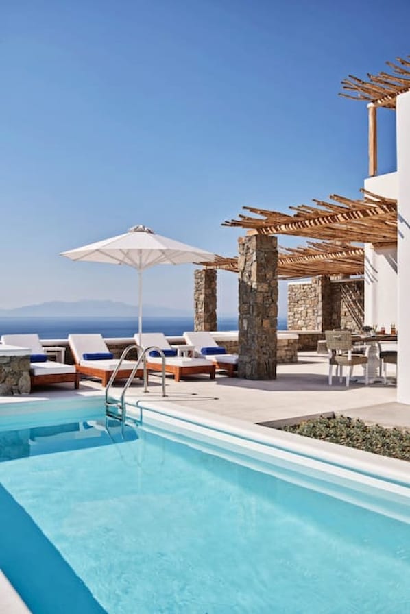 Un espace piscine doté de chaises longues et de parasols, de piliers en pierre et offrant une vue sur la mer.