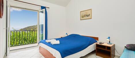 Een slaapkamer met een bed, nachtkastje, lamp, foto aan de muur en balkon met uitzicht op groen en heuvels.