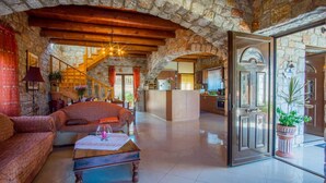 A spacious living area with a stone wall, wooden beams, a sofa, a coffee table, and a kitchen in the background. - Villa Vicina - In Platanias and 4min by the beach (GV2W+PVV Platanias, Greece)