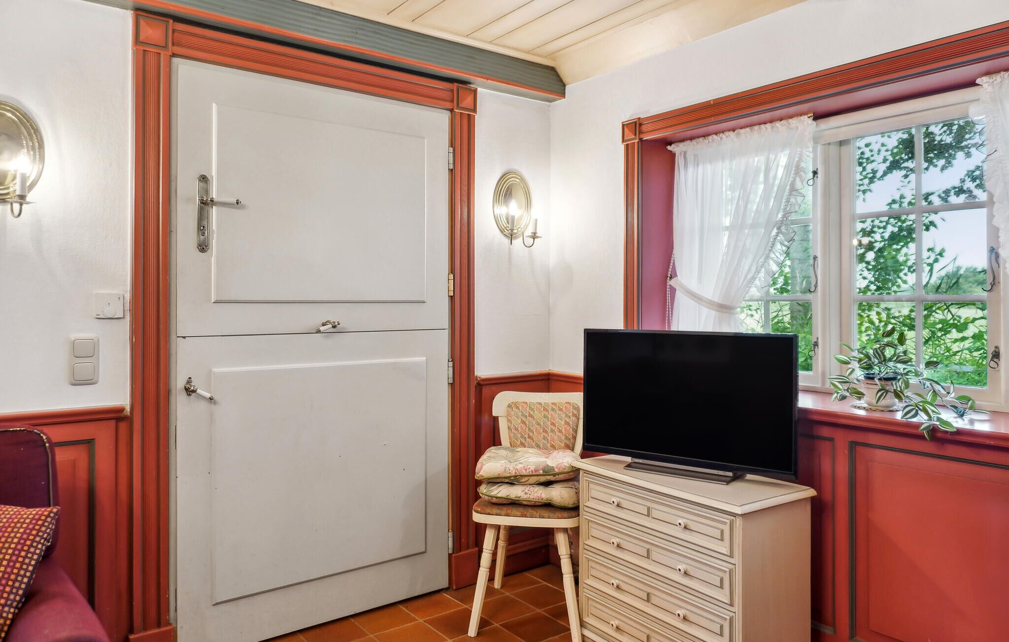 Ein Zimmer mit geschlossener Tür, einem Fernseher auf einem Schrank, einem Sessel und einem Fenster mit Vorhängen.