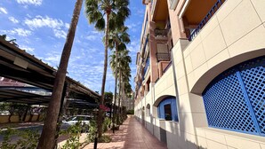 A corridor lined with palm trees and a covered walkway, leading to a building with blue lattice windows and a tiled facade. - Vilamar-1-d  VYB Sea Views (Denia)