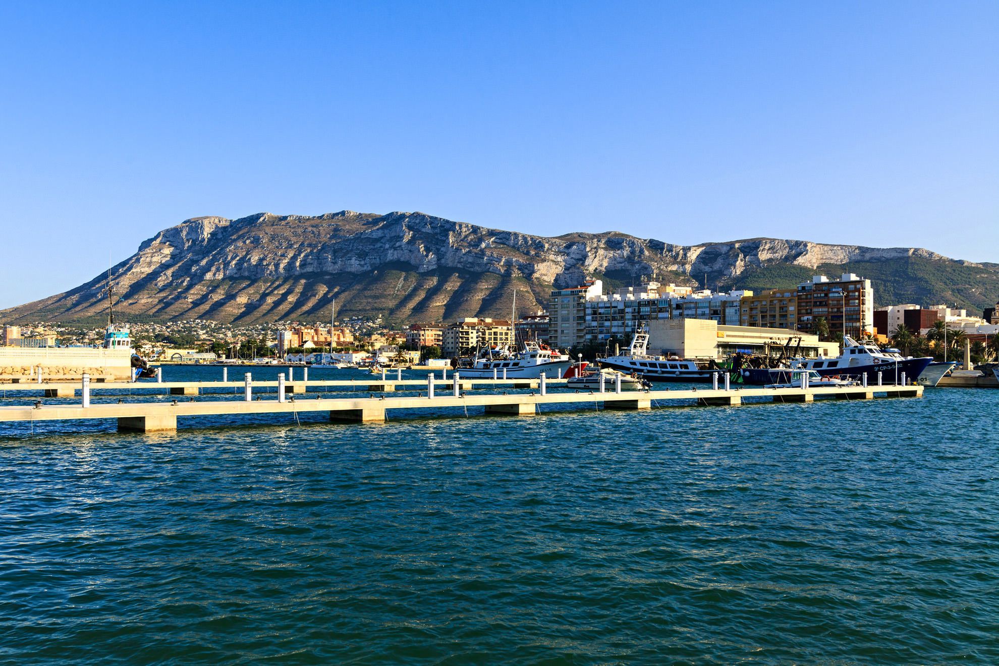 Un port de plaisance sur la côte, avec des bateaux amarrés, une jetée s’avançant dans l’eau et une chaîne de montagnes en arrière-plan.