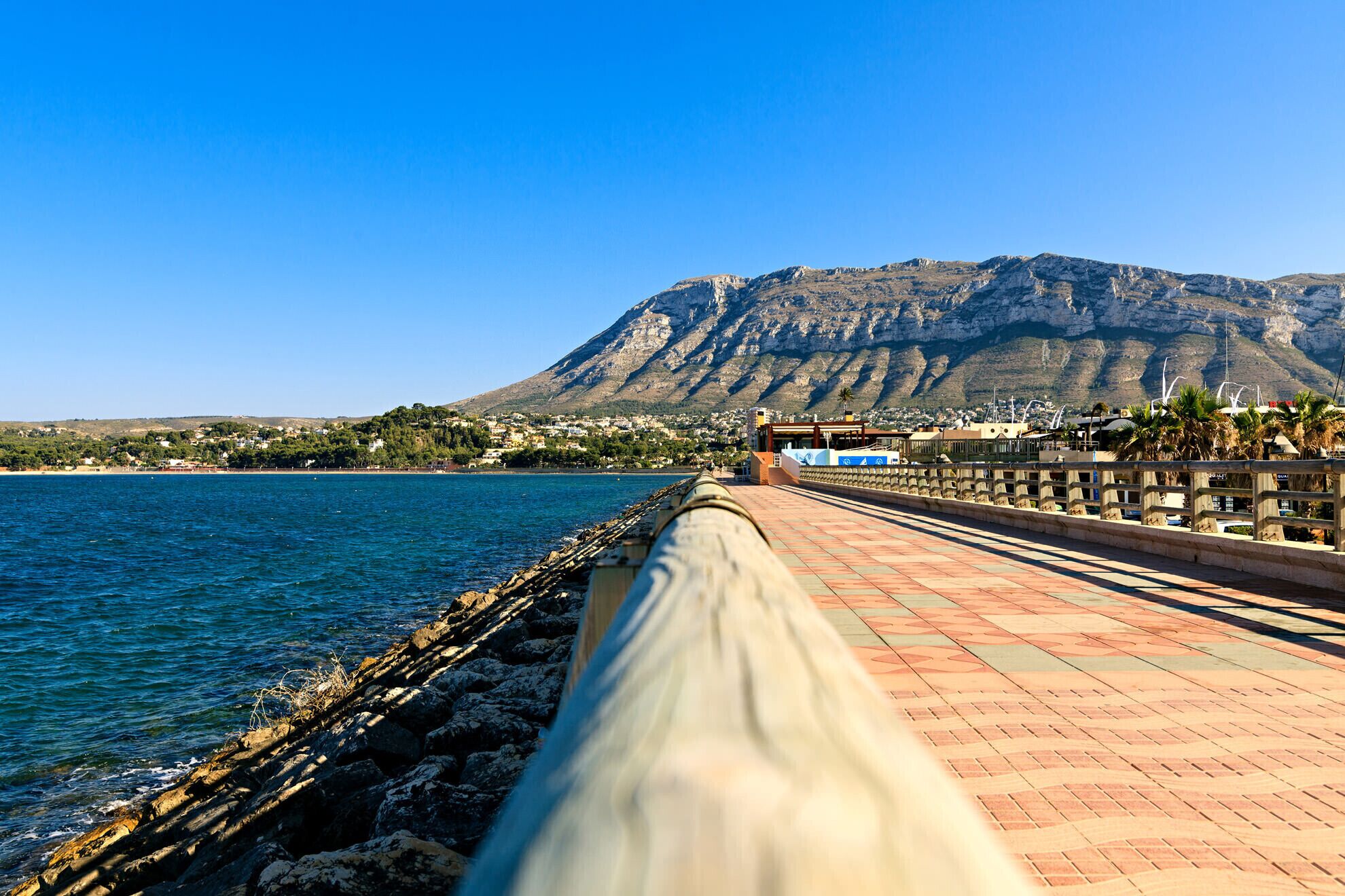 Eine Uferpromenade mit Blick auf ein Gebirge und eine Stadt in der Ferne.
