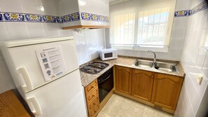 A kitchen with white tiles, a refrigerator, a microwave, a stove, wooden cabinets, and a window with a view.