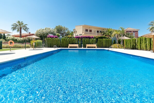 A clear blue swimming pool with lounge chairs, surrounded by neatly trimmed hedges and a hotel named 'Hotel Rosa' in the background.