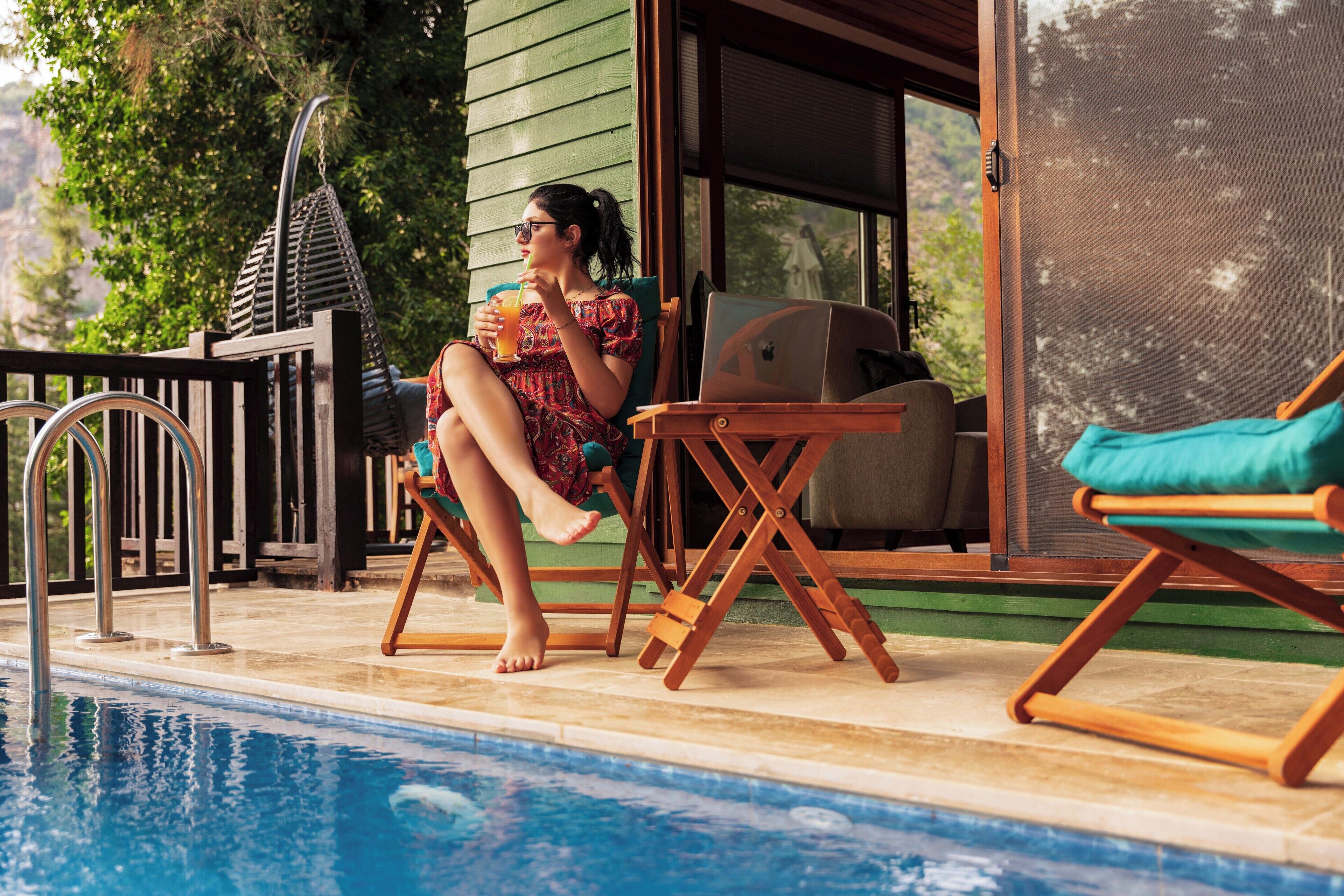 Eine Person entspannt in einer Liege am Pool, mit einem Laptop und einem Grill, in der Nähe eines Pools mit Holzterrasse und einer Hängematte.