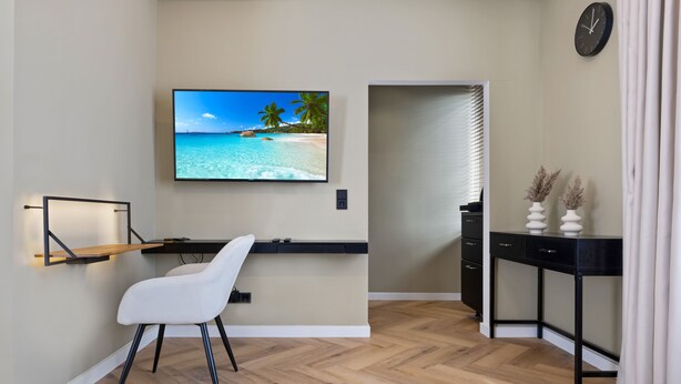 Une chambre moderne avec un parquet en bois disposé en motif chevrons, une chaise blanche, un bureau avec un téléviseur à écran plat, une horloge noire et une petite plante posée sur une table d’appoint.