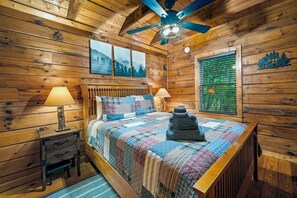 Un dormitorio de cabaña de madera con una cama, dos lámparas de noche, un ventilador de techo y una ventana con vistas a árboles.