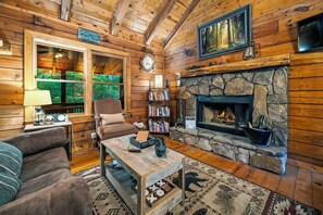 Interior acogedor de una cabaña con chimenea de piedra, paredes de madera, una estantería y un ventanal con vistas a la vegetación.