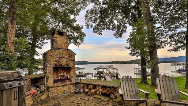 An outdoor stone fireplace with a grill, surrounded by Adirondack chairs and a dock extending into a lake. - A Lake Blue Ridge Cottage | Morganton, GA (Morgantown)
