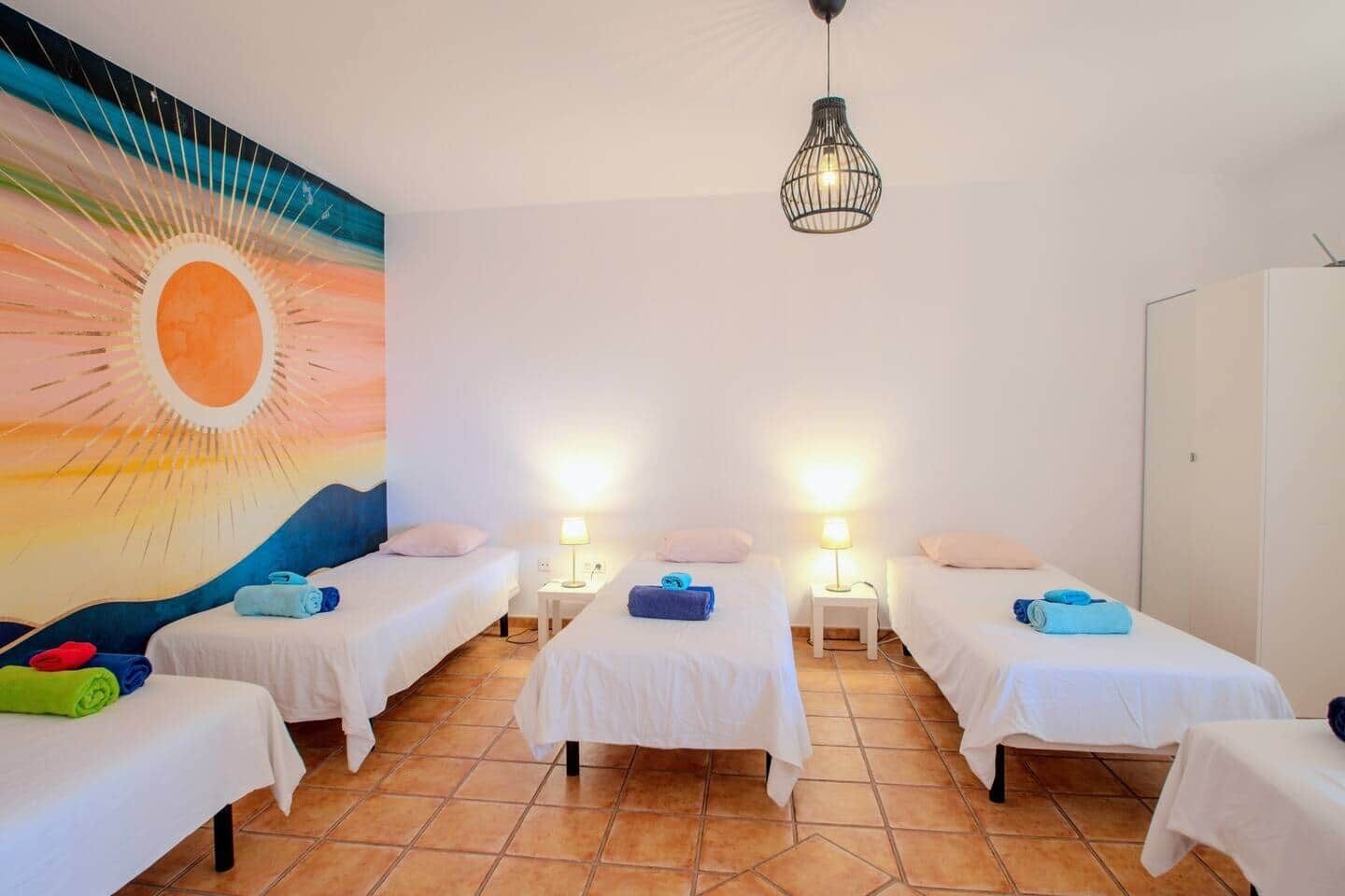 A hotel room with four single beds, each with a white bedspread and a small bedside table with a lamp. There is a large mural on the wall depicting a sunset.