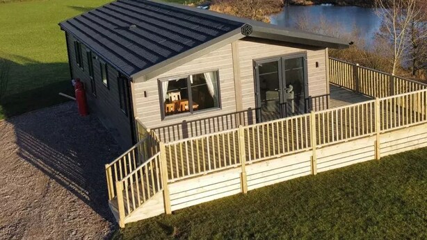 A modern cabin with a deck overlooking a lake, surrounded by rolling hills and greenery. - Luxury 3 bedroom, pet-friendly lodge on the side of a beautiful fishing lake. (Clitheroe)