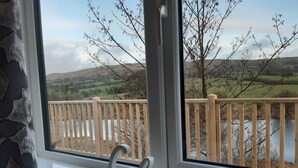 A window with a view of a wooden deck overlooking a river and hills. - Luxury 3 bedroom, pet-friendly lodge on the side of a beautiful fishing lake. (Clitheroe)