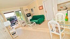 Un espacio habitable compacto con un sofá verde, una mesa de vidrio, sillas blancas, un televisor y una zona de cocina.