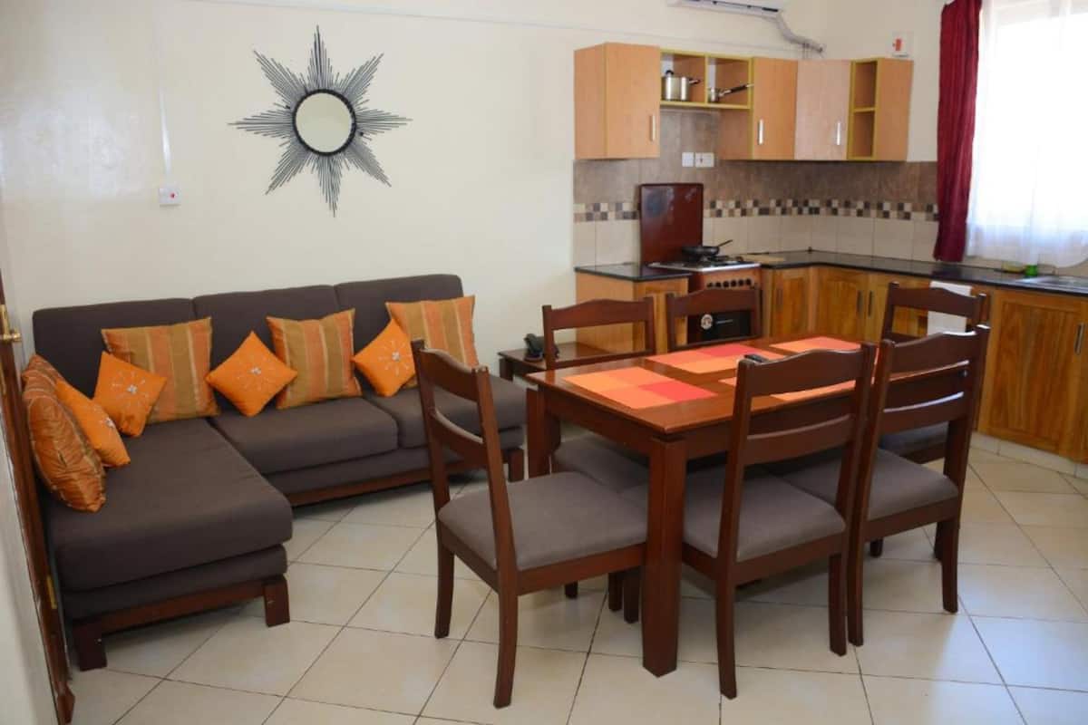 A living room with a sofa, chairs, and a dining table. There is a kitchen in the background with wooden cabinets and a stove.