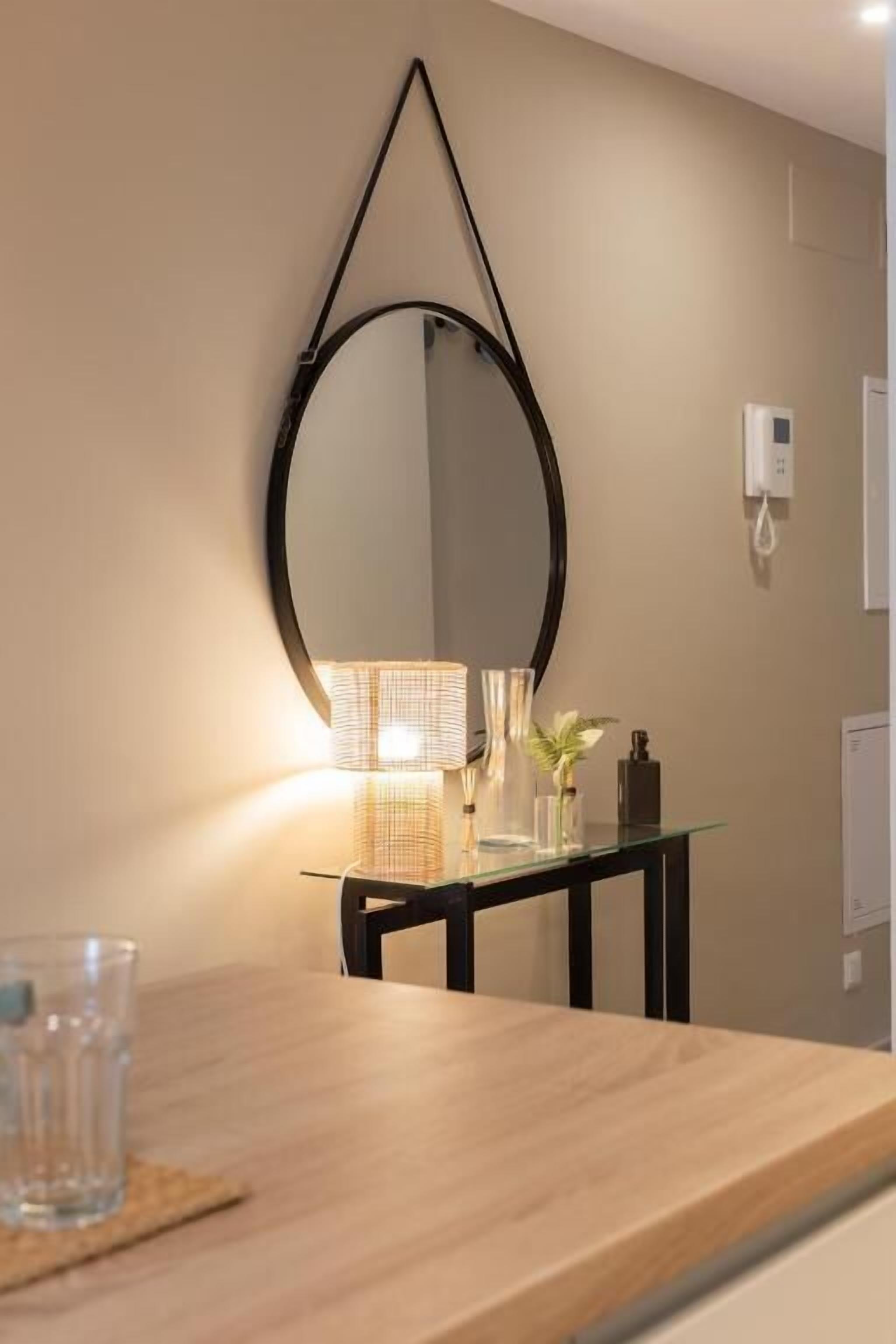 A modern bathroom with a wooden vanity, a round mirror, and a glass table with a lamp and decorative items.
