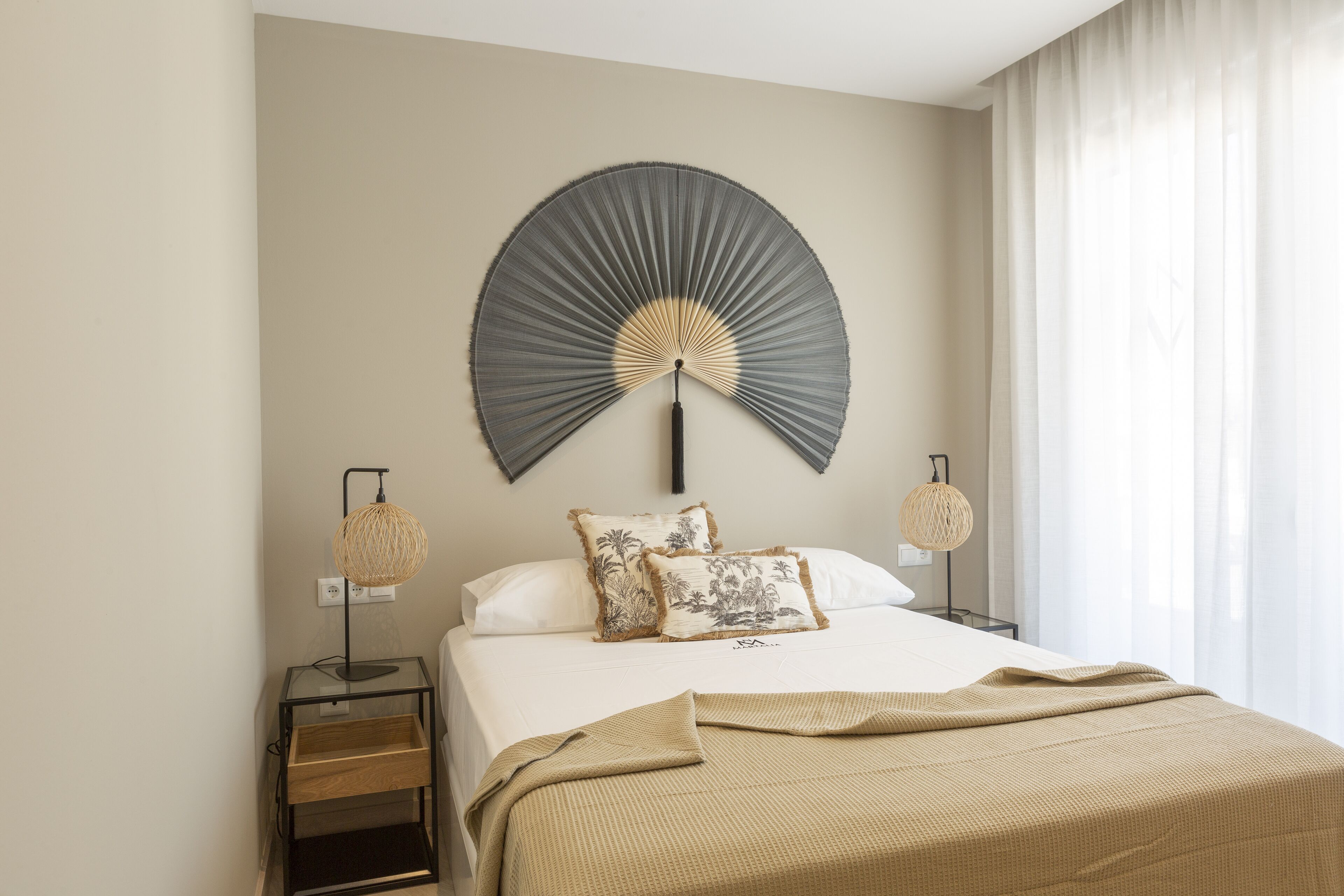 Un lit bien fait, recouvert d’une couverture beige, avec deux lampes de chevet et un ventilateur fixé au mur.