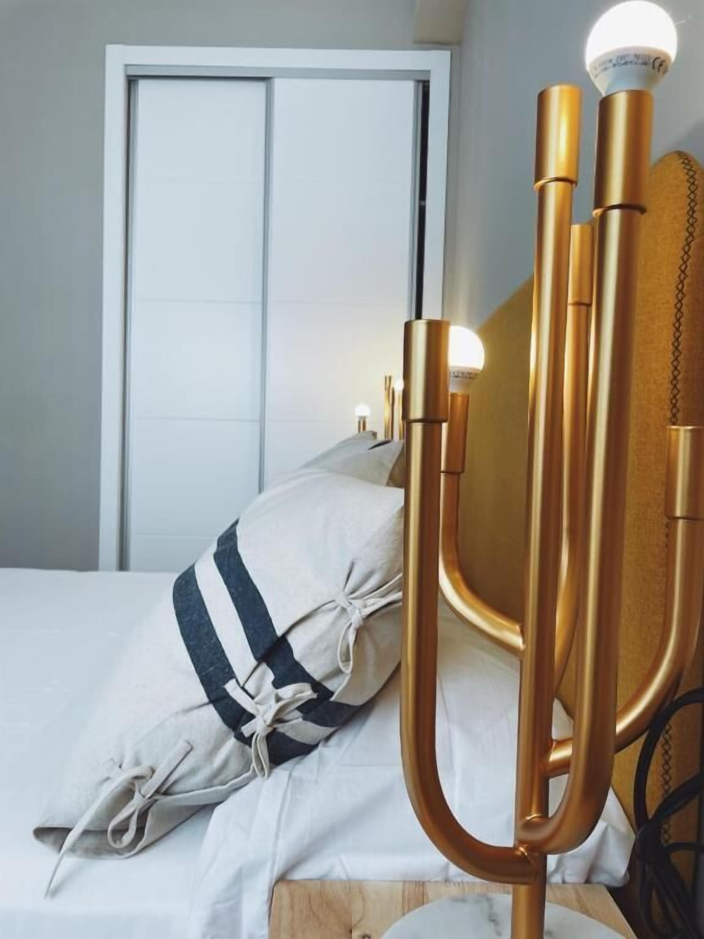 Una cama con un cojín a rayas blanco y azul, un candelabro dorado con una bombilla encendida, y una puerta blanca al fondo.