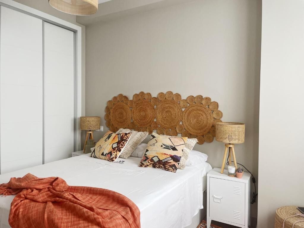 Una cama bien hecha con almohadones estampados, un cabecero tejido, lámparas de noche y una mesita con una planta.