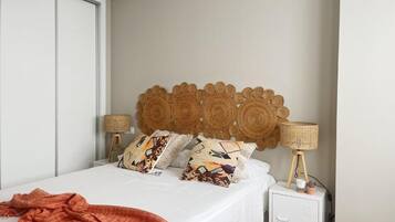 Una cama bien hecha con almohadones estampados, un cabecero tejido, lámparas de noche y una mesita con una planta.