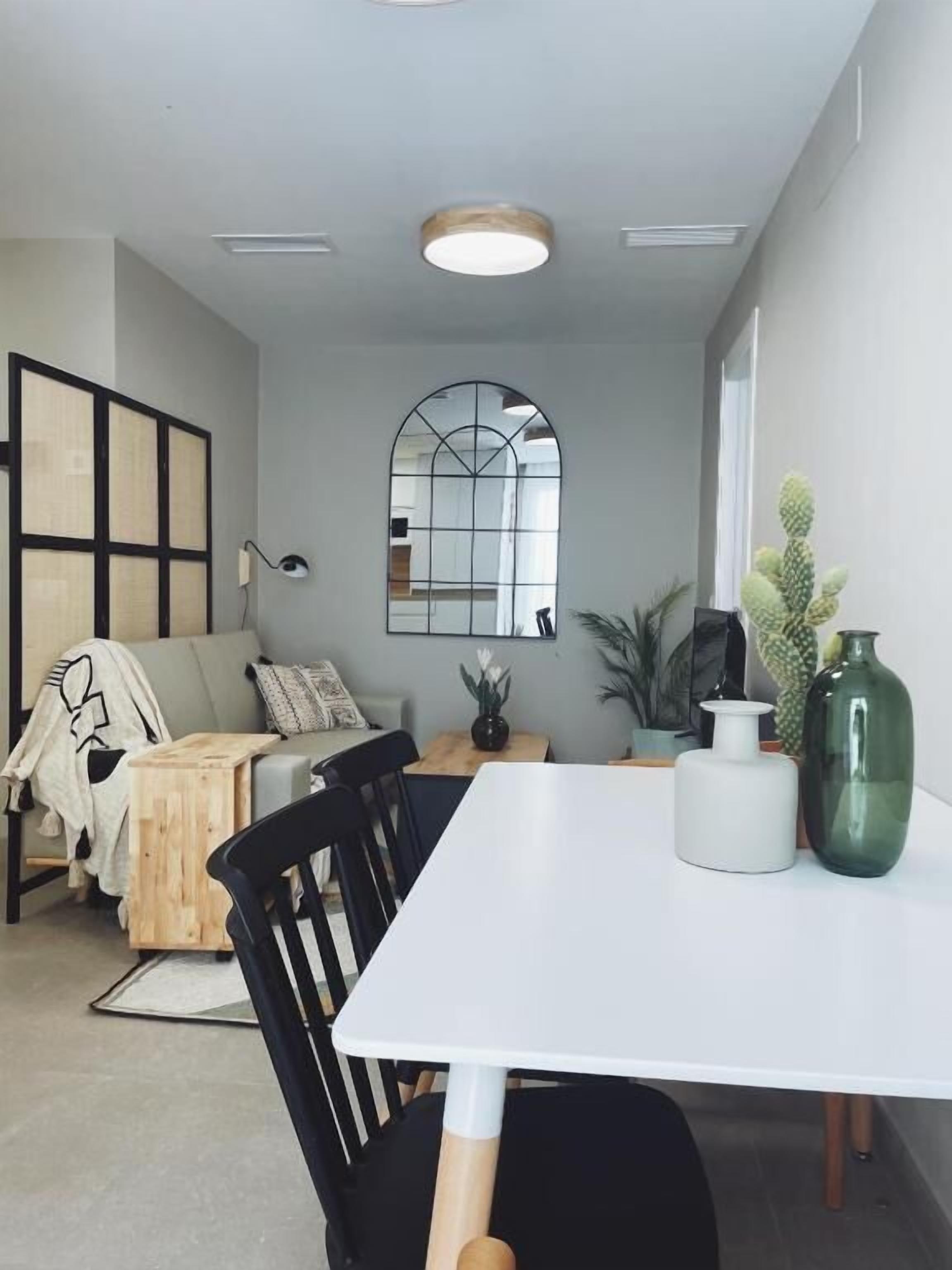 A modern dining area with a white table, black chairs, a wooden side table, and a large window with a geometric mirror.