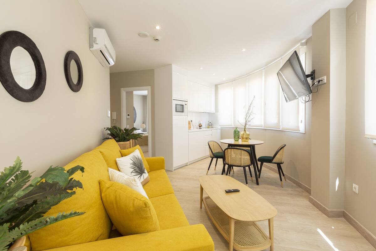 Un salón luminoso con un sofá amarillo, una mesa de centro de madera y un comedor con una mesa pequeña y sillas.