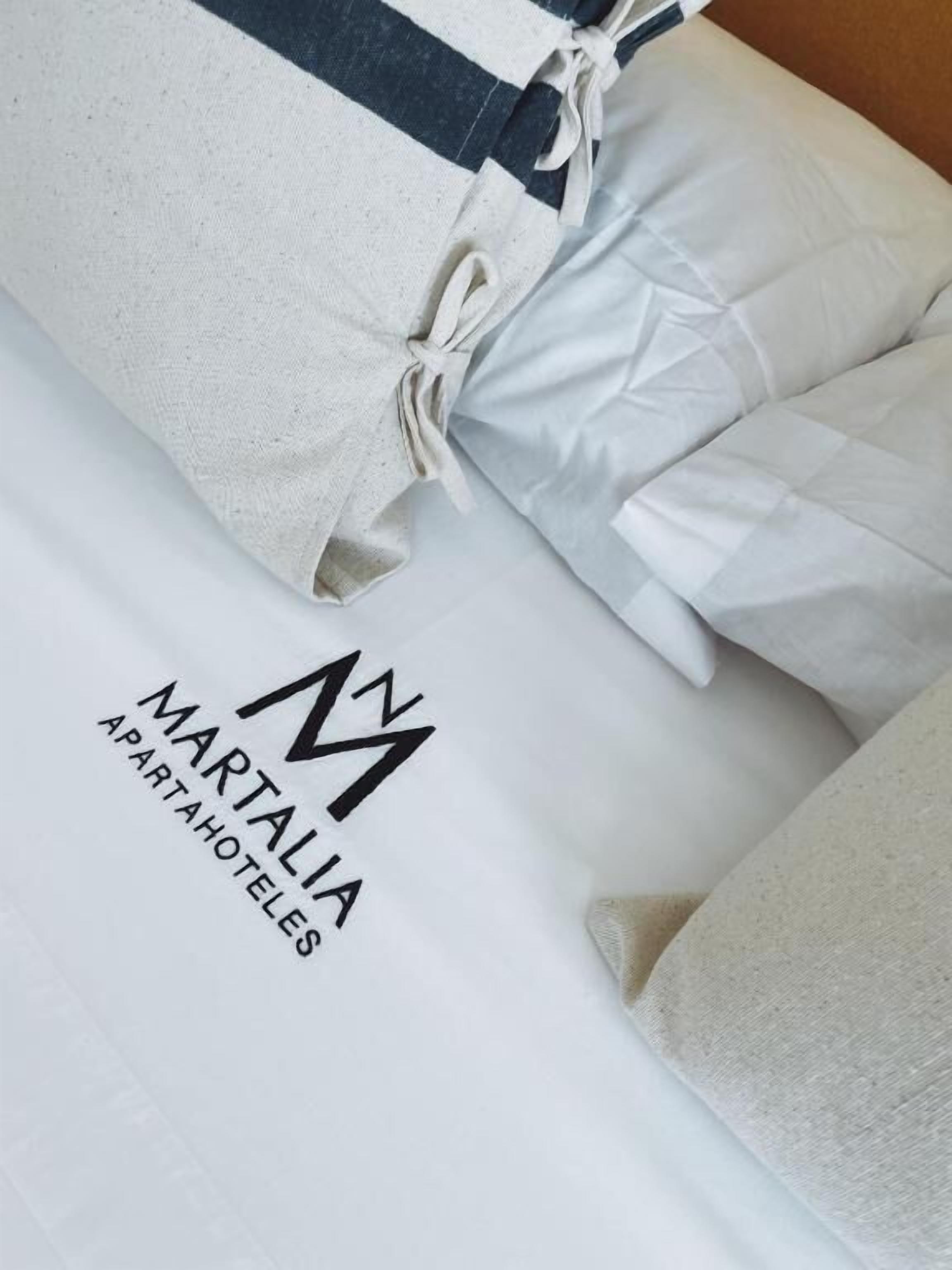 Valkoisilla lakanoiden ja tyynyjen peitetty sänky, ja siihen kiinnitetty etiketti, jossa lukee: ”MARTALIA APARTAHOTELES”.