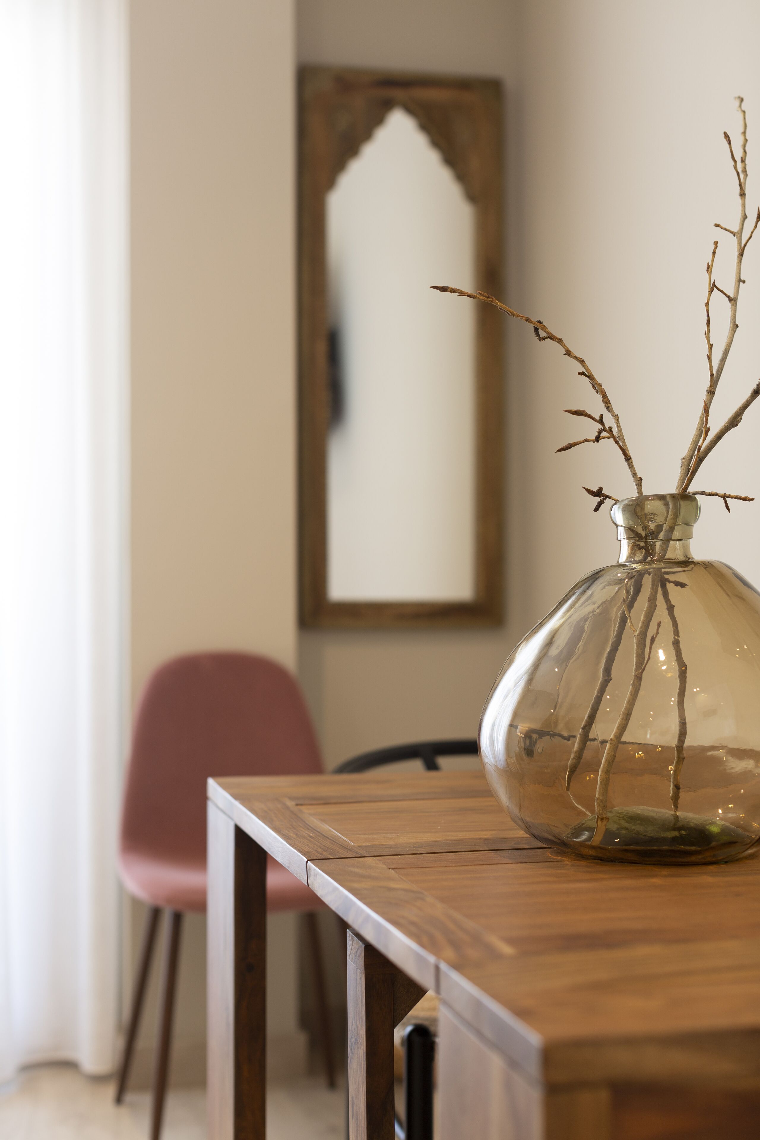 Una mesa de madera con un florero de vidrio transparente que contiene ramas, una silla roja y un espejo enmarcado en la pared.