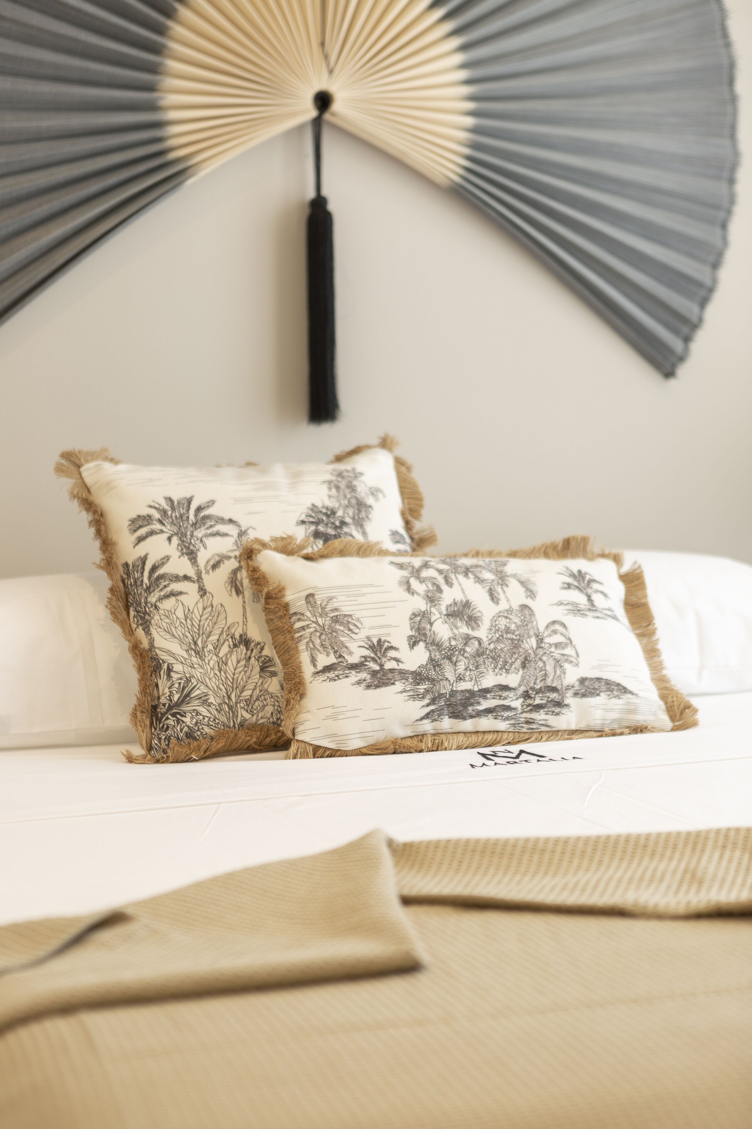 A bed with two decorative pillows featuring palm tree designs, a black tassel hanging from a fan, and a beige throw blanket.