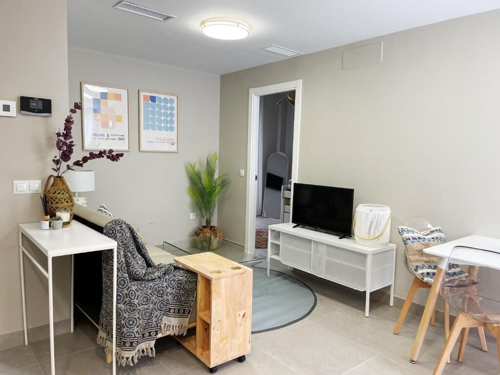 Una sala de estar moderna con televisor, muebles blancos, una mesita de madera y una planta.