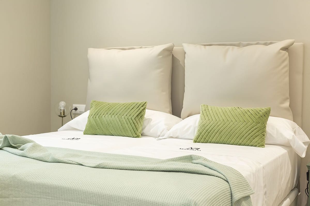 Una cama bien hecha con ropa de cama blanca y verde, una mesita de noche con una lámpara y un enchufe de pared.
