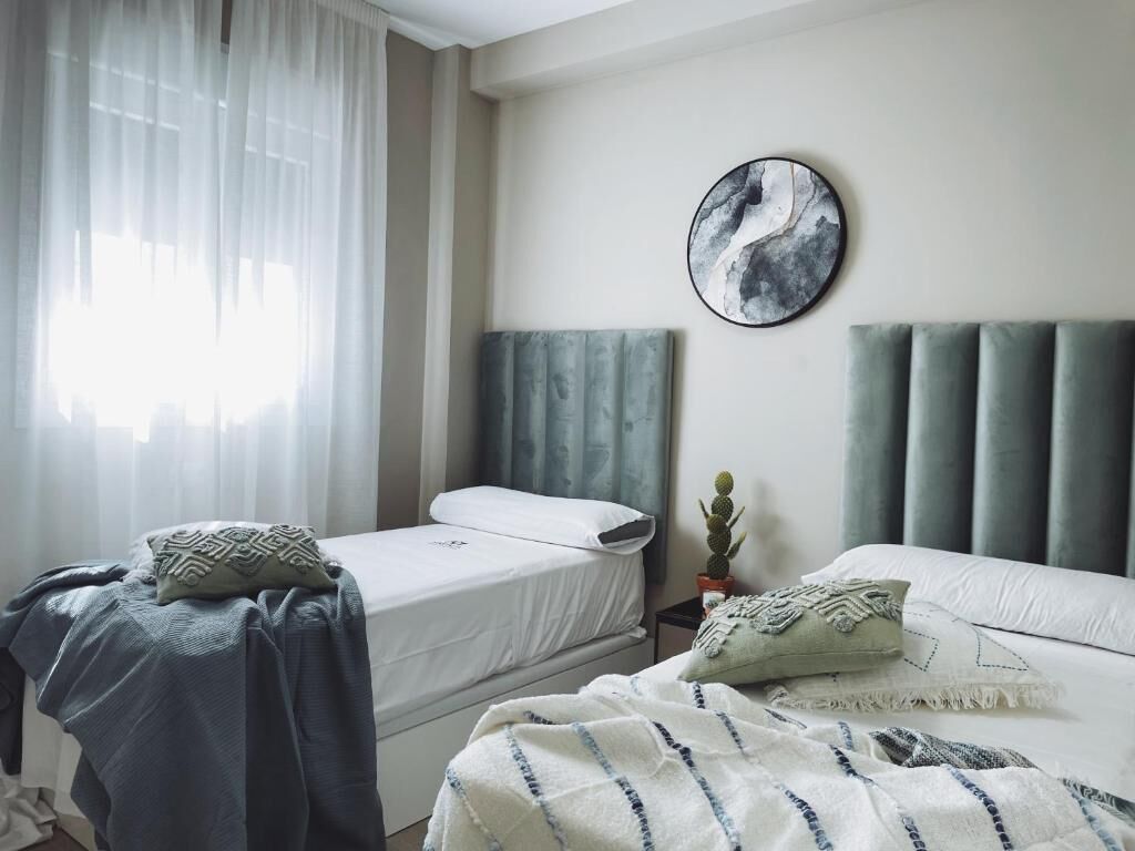 Un dormitorio con dos camas, un ventanal con cortinas translúcidas, una obra de arte circular en la pared y un cactus sobre una mesita de noche.