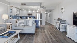 Een moderne woonkamer met een grijze bank, een salontafel en een tv-meubel. Er is een keuken met witte kasten en een blauwe achterwand.