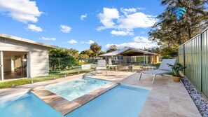 A modern outdoor pool area with a hot tub, lounge chairs, and a glass-enclosed building.