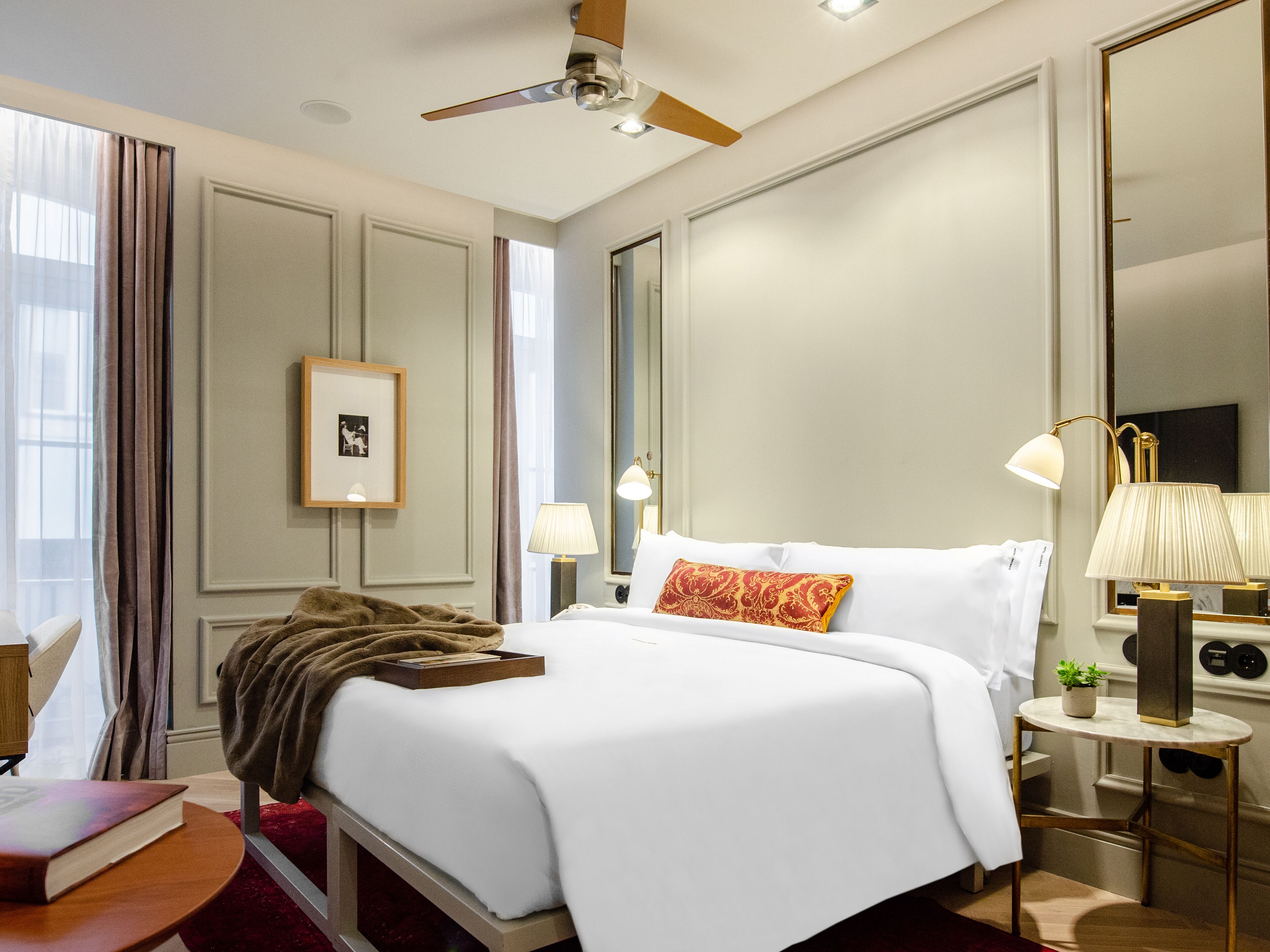 Une chambre d’hôtel avec un lit, des lampes de chevet, un ventilateur de plafond, un tableau encadré au mur et une petite table sur laquelle se trouvent un livre et une plante.