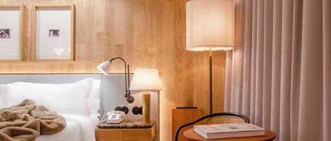 A hotel room with a bed, bedside table with a lamp, a small table with a chair, and a large window with curtains.