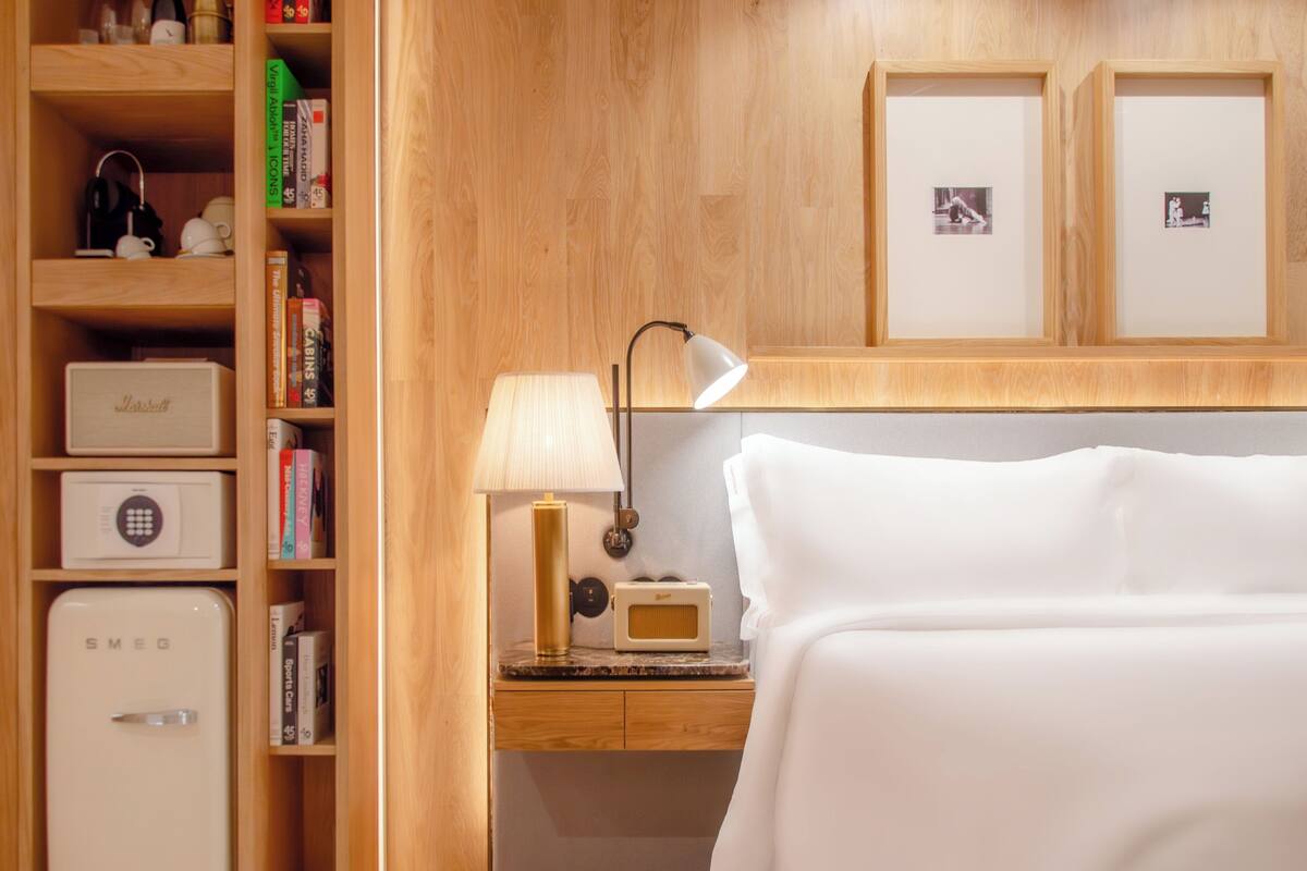 Ein Hotelzimmer mit einem Bett, einem Nachttisch mit Lampe, einem Holzregal und einem kleinen Kühlschrank.